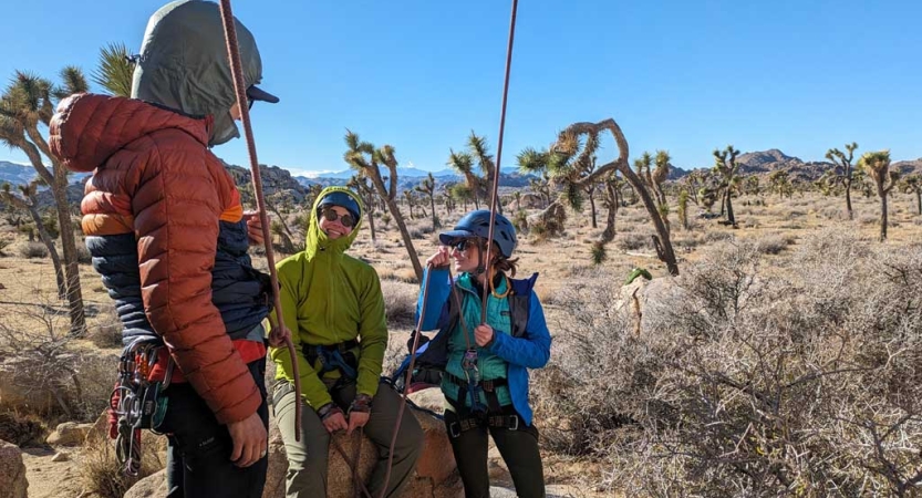 Three people wearing safety gear stand in a desert environment. Two of the people are attached to ropes that appear taut. 